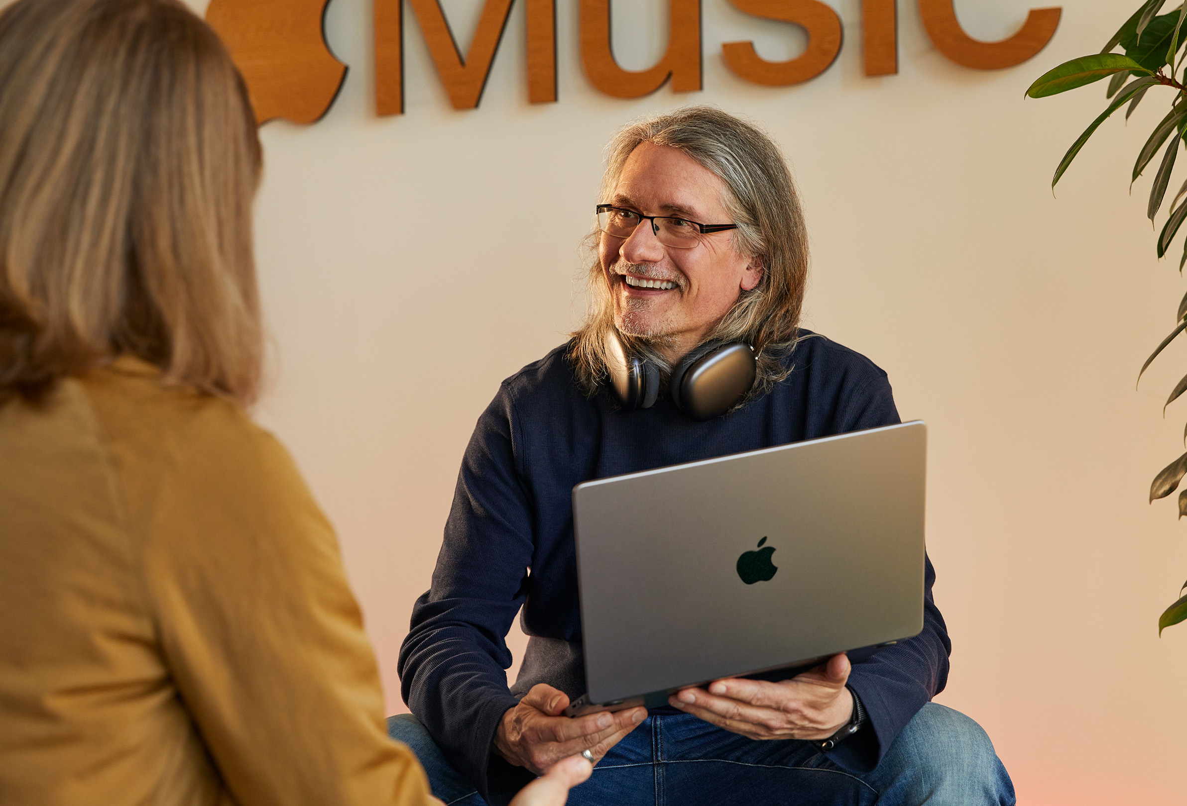 Two Apple employees in conversation, one holding a MacBook and sitting in front of a wall with the Apple?Music logo on it.