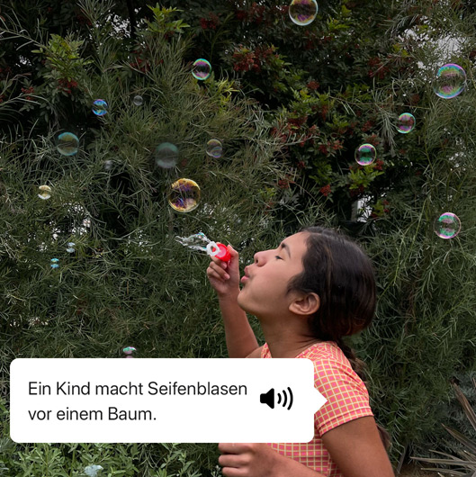 VoiceOver beschreibt ein Foto und zeigt die Sprachausgabe. ?Ein Kind macht Seifenblasen vor einem Baum.“