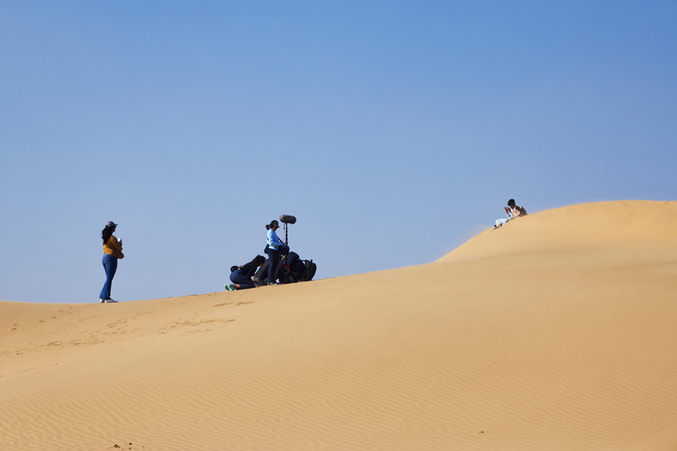 A film crew is shown working in a desert with an actor sitting alone on a dune on the set of “Mirage.”