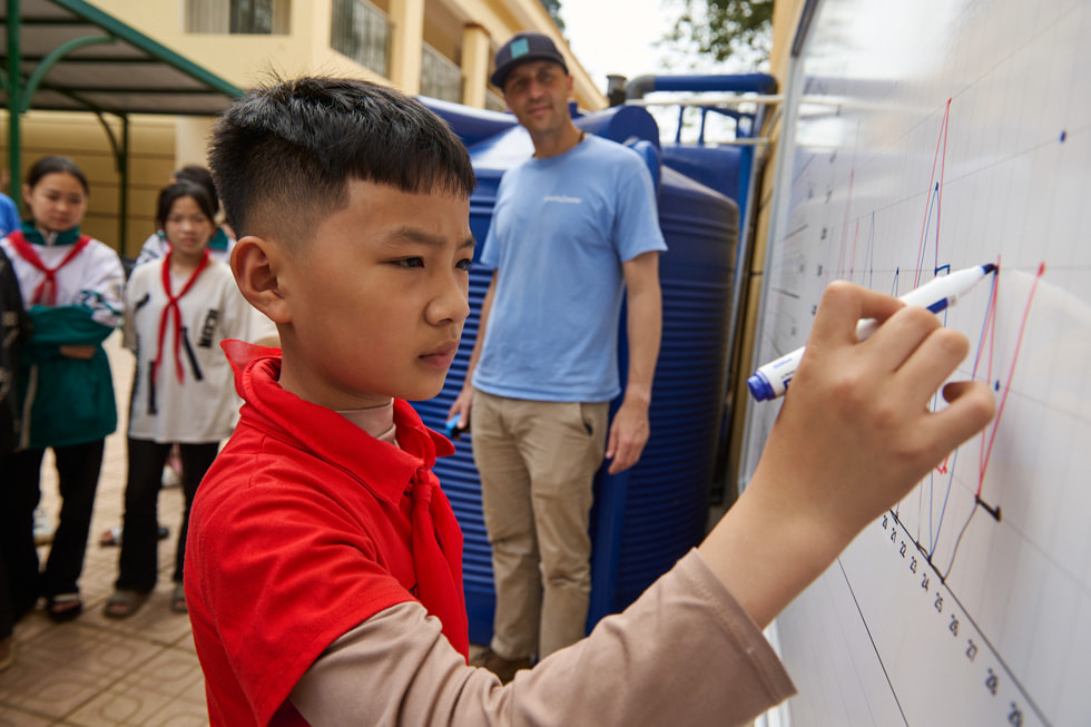 A student writes on a dry-erase board as fellow students and adults look on.